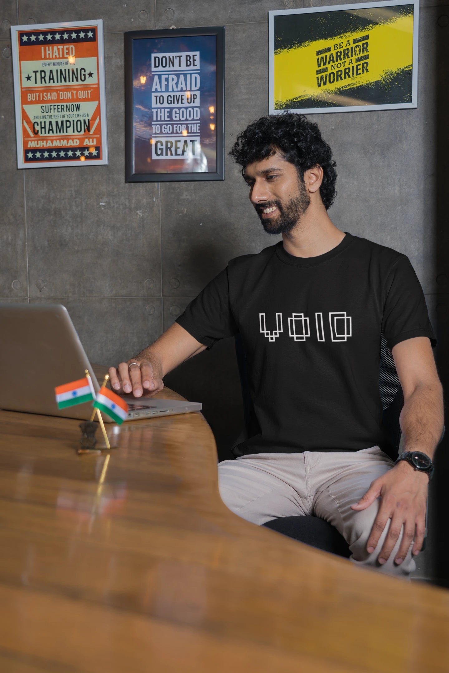 Man sitting at a desk with a laptop, wearing a black t-shirt with 'VOIO' branding, in an office setting.