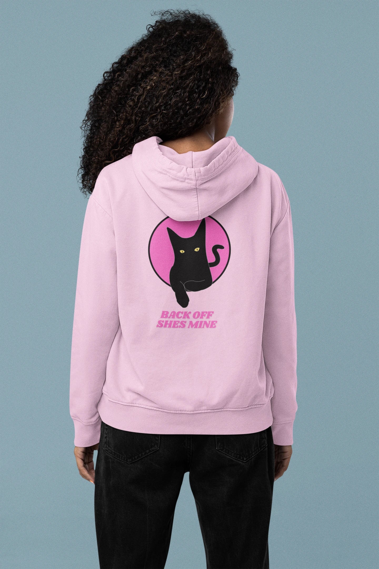 Person wearing a pink hoodie with a black cat graphic and text on the back against a gray background