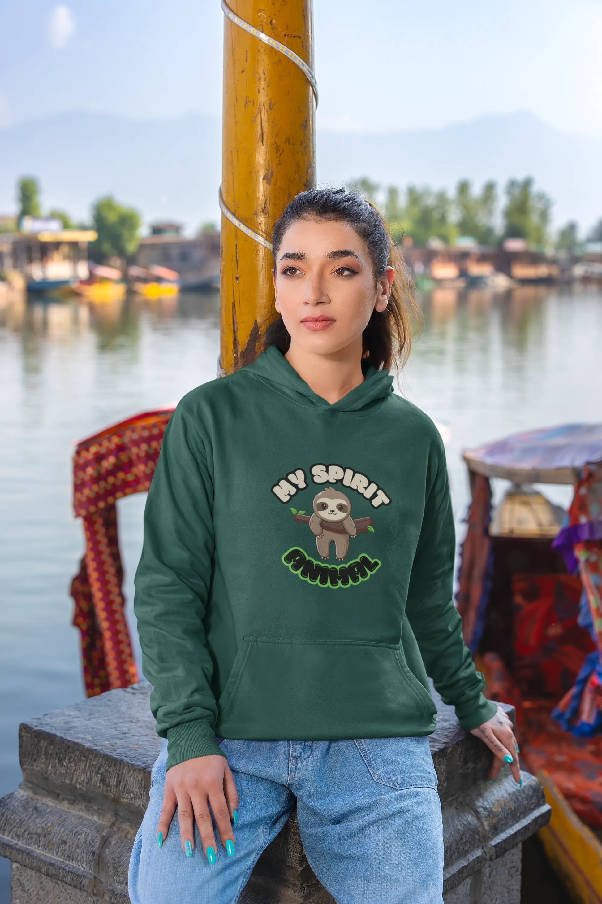 Woman wearing a green hoodie with a sloth graphic and text by a waterfront.
