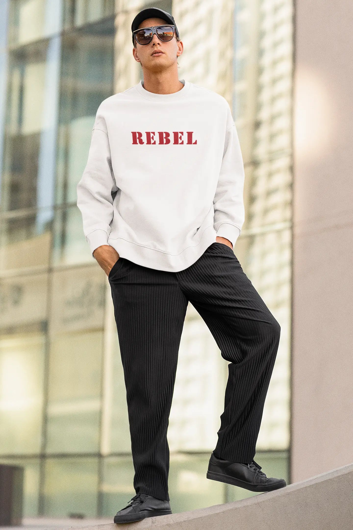 Person wearing a white sweatshirt with 'REBEL' printed in red, black pants, and black shoes, standing against a modern building.