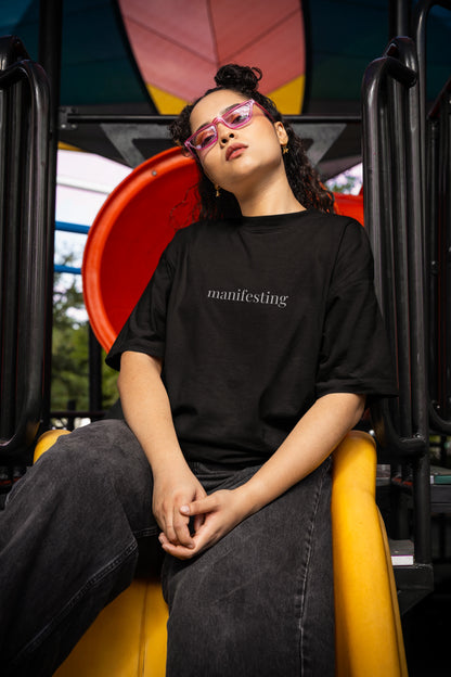 Person wearing a black t-shirt with 'manifesting' text, sitting on a colorful playground structure.