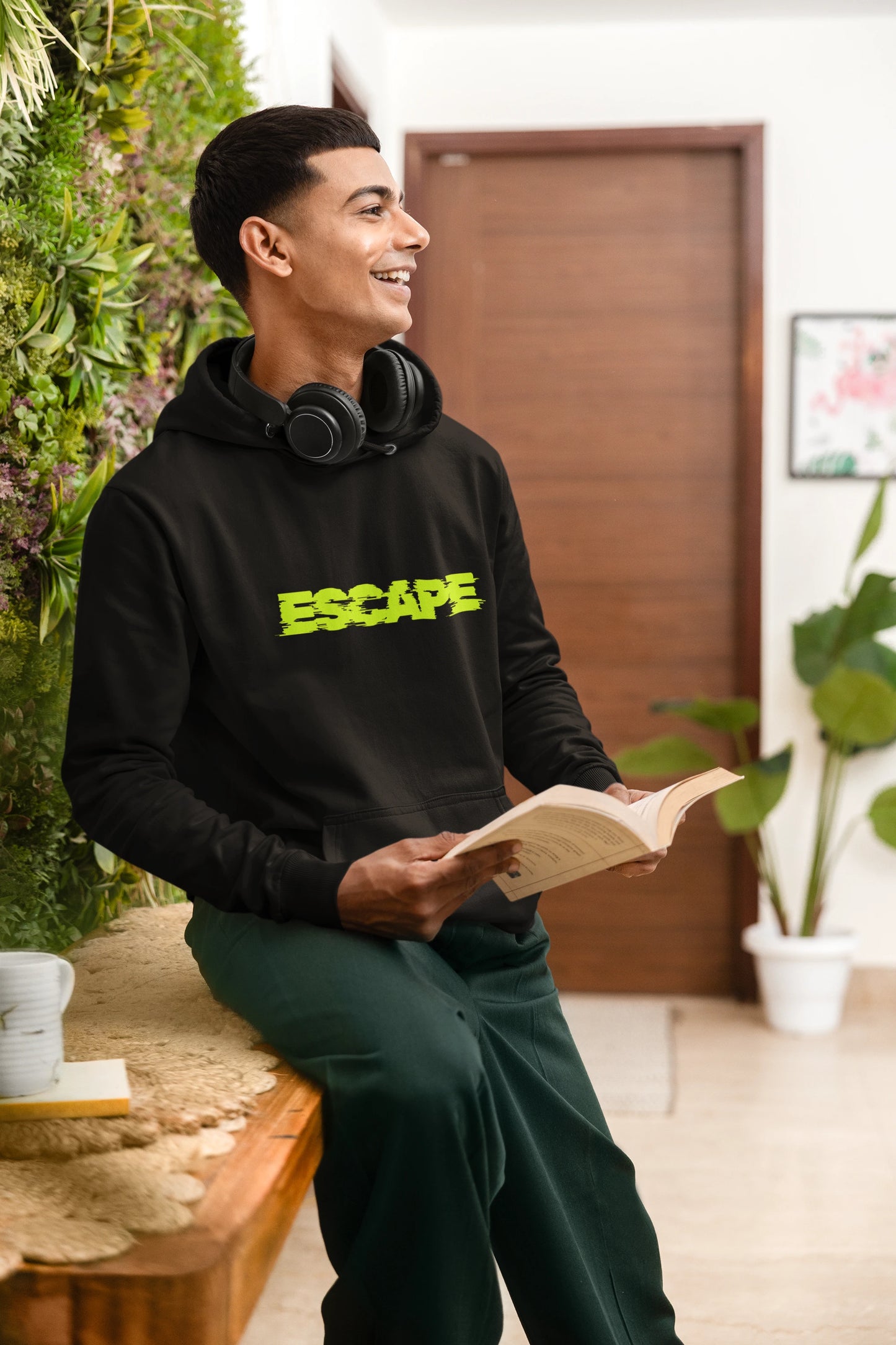 Man wearing a black hoodie with 'ESCAPE' in yellow, sitting and reading a book in a room with plants.